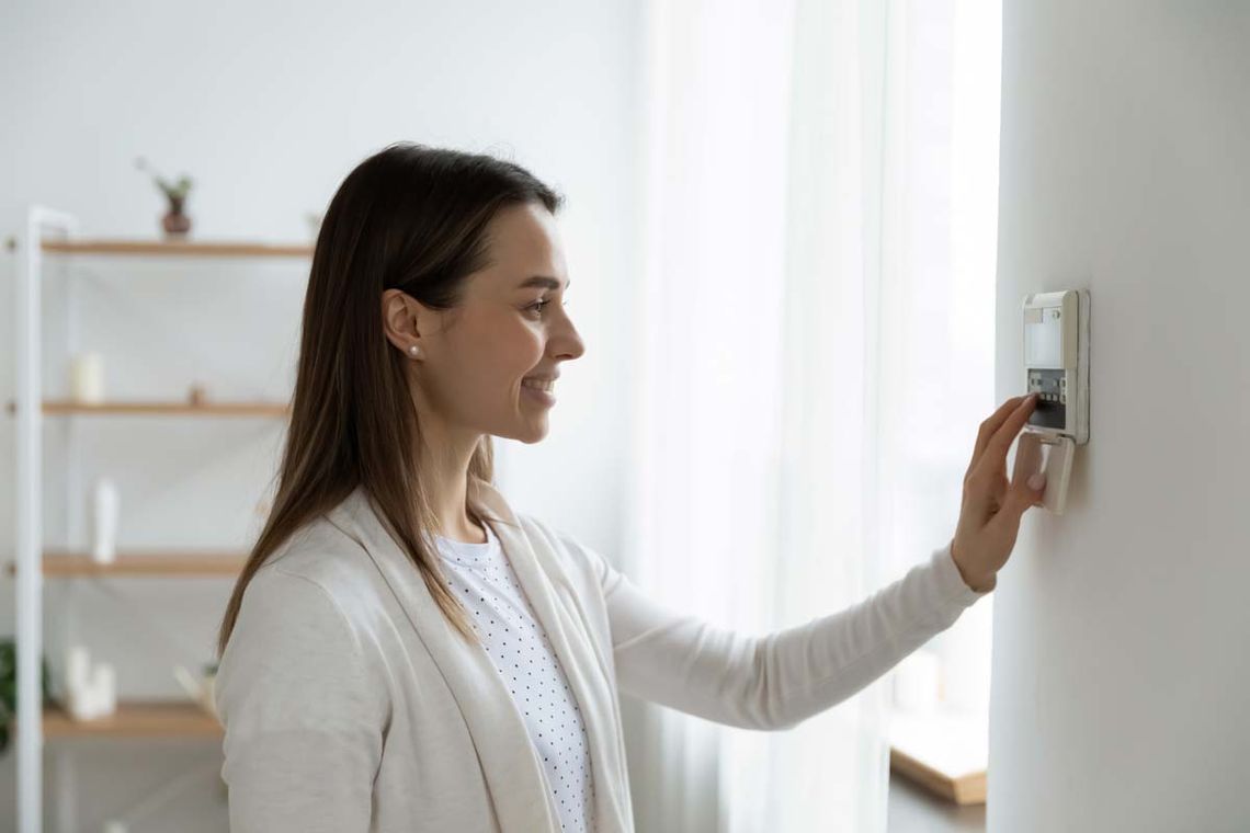 Smiling woman standing near wall-mounted device adjusting degrees in living room set comfortable temperature using thermostat home heating system. 