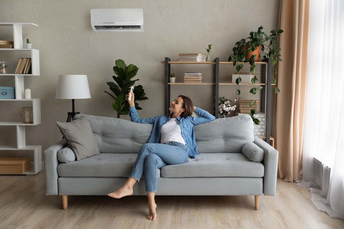 Happy young Caucasian woman relax on couch in living room turn on air conditioner with remote controller